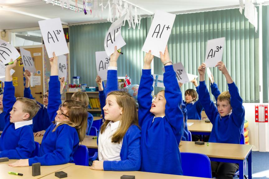 Classroom of children with their hands up and boards in the air. 